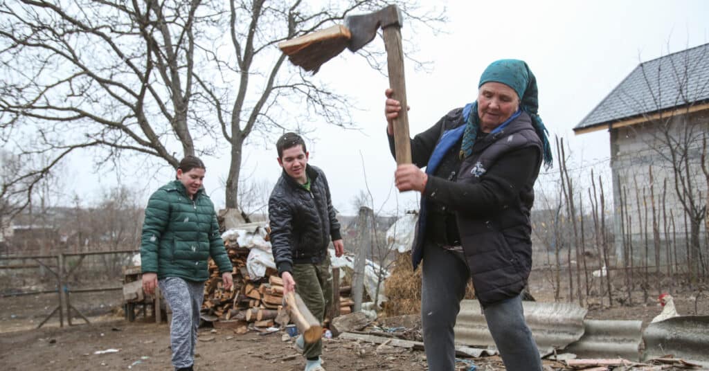 An elderly woman wearing a turquoise headscarf chops wood, while her young adult grandchildren stand to the side, helping her.