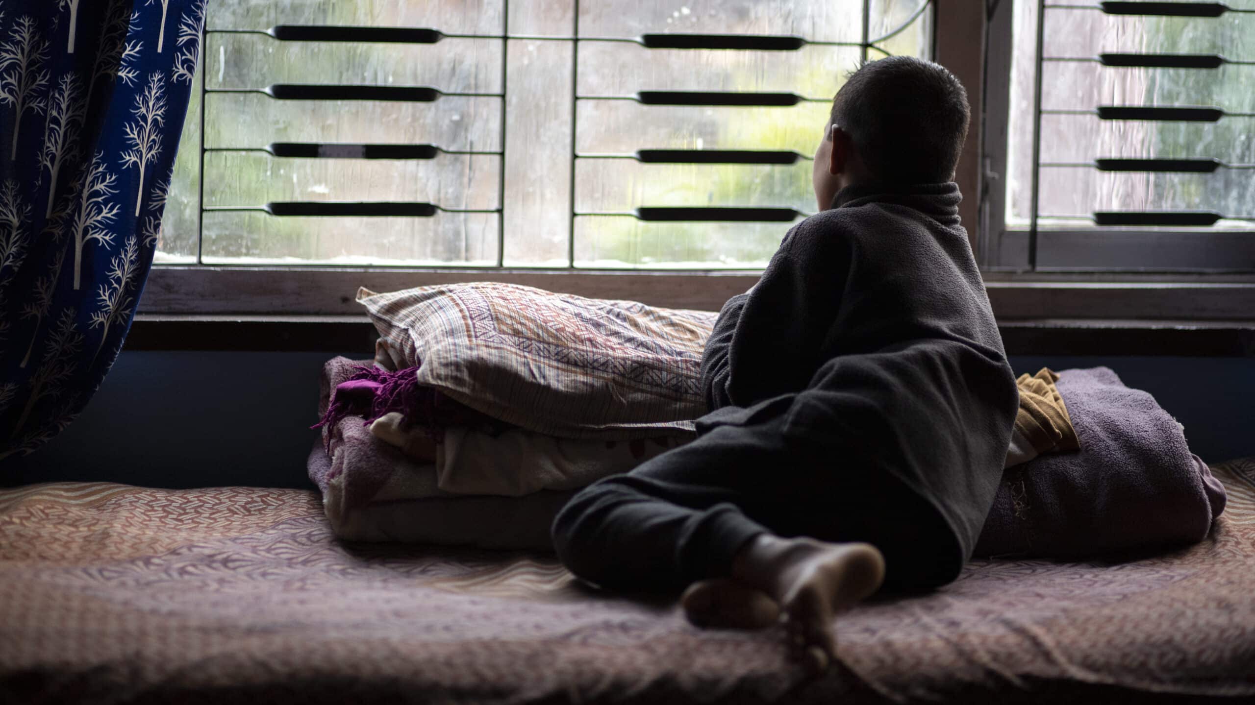 A child with their back turned gazes sadly out of the barred windows of an orphanage
