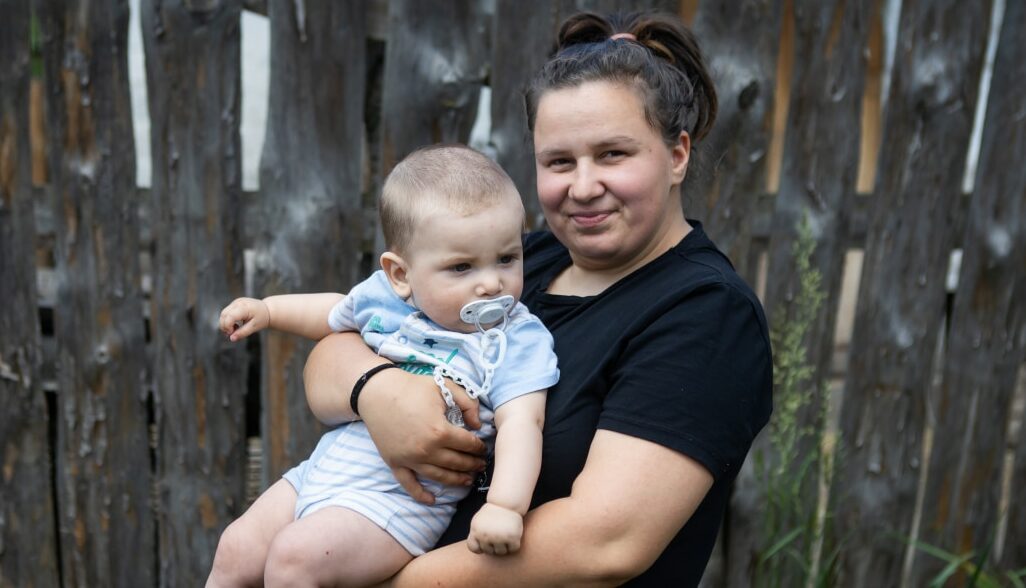 Young brunette mum from Ukraine smiles proudly while holding her baby boy in her arms.