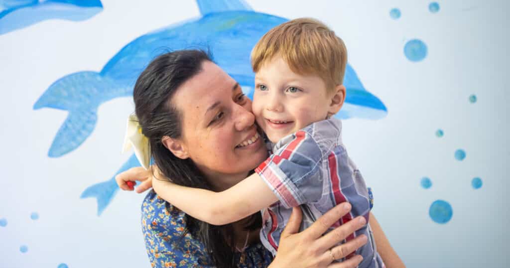 Olga holds her son, Nikita. He's wearing a checkered shirt, smiling as his mum hugs him close.
