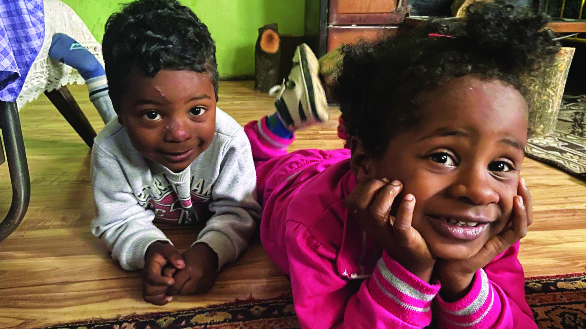 A young boy and girl lie on their front on the floor of their house, chins in hand looking up at the camera smiling