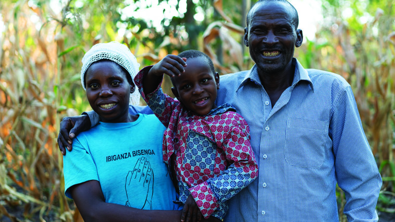A Rwandan boy, Carrol, is held by his mother, as his father wraps his arm around her shoulder.