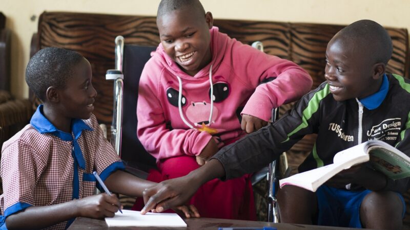 Cary sits in a wheelchair between two other children, one holds a textbook and the other holds a pen to a notepad