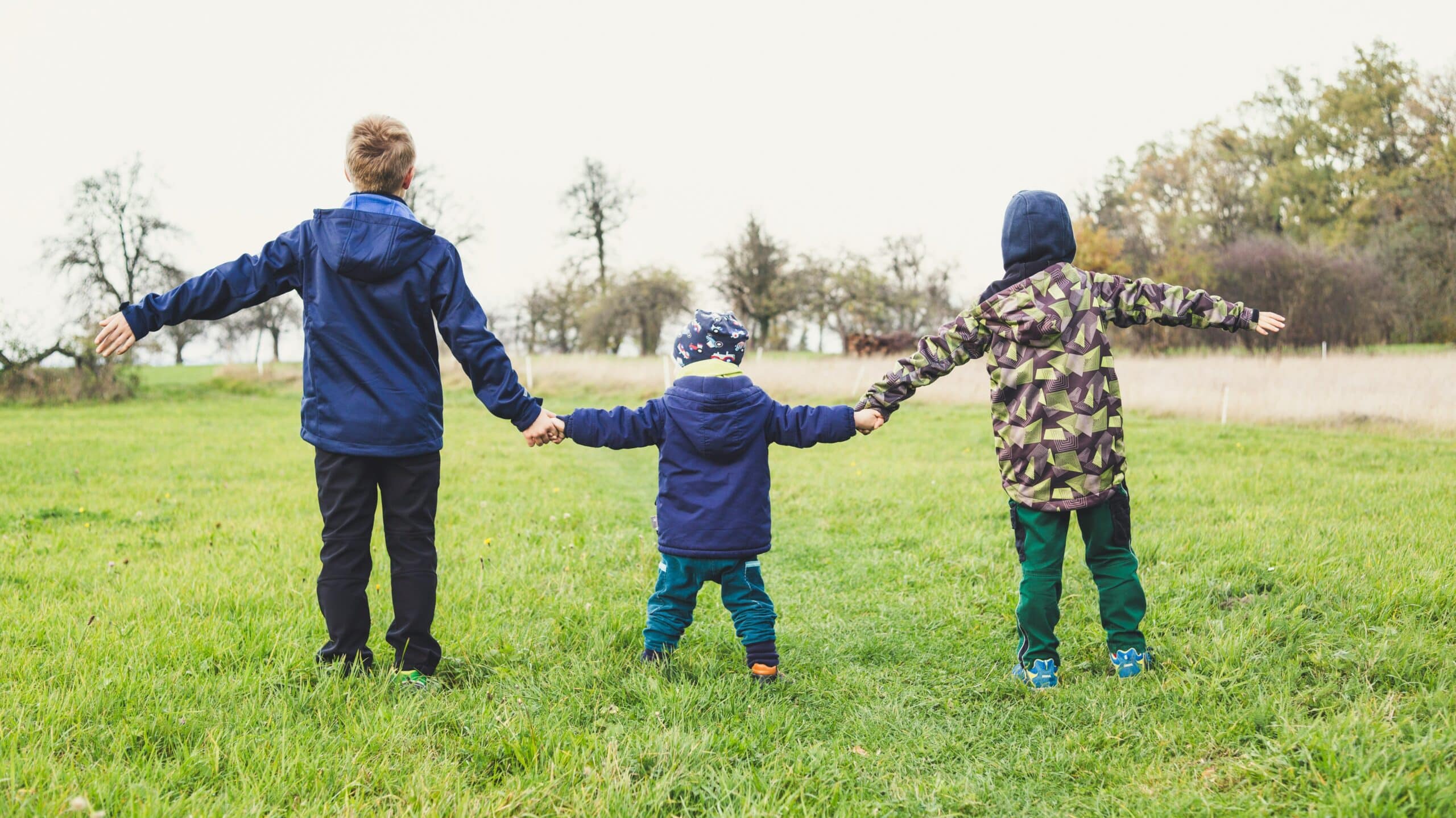 Three small children hold hands and walk away into a green field.