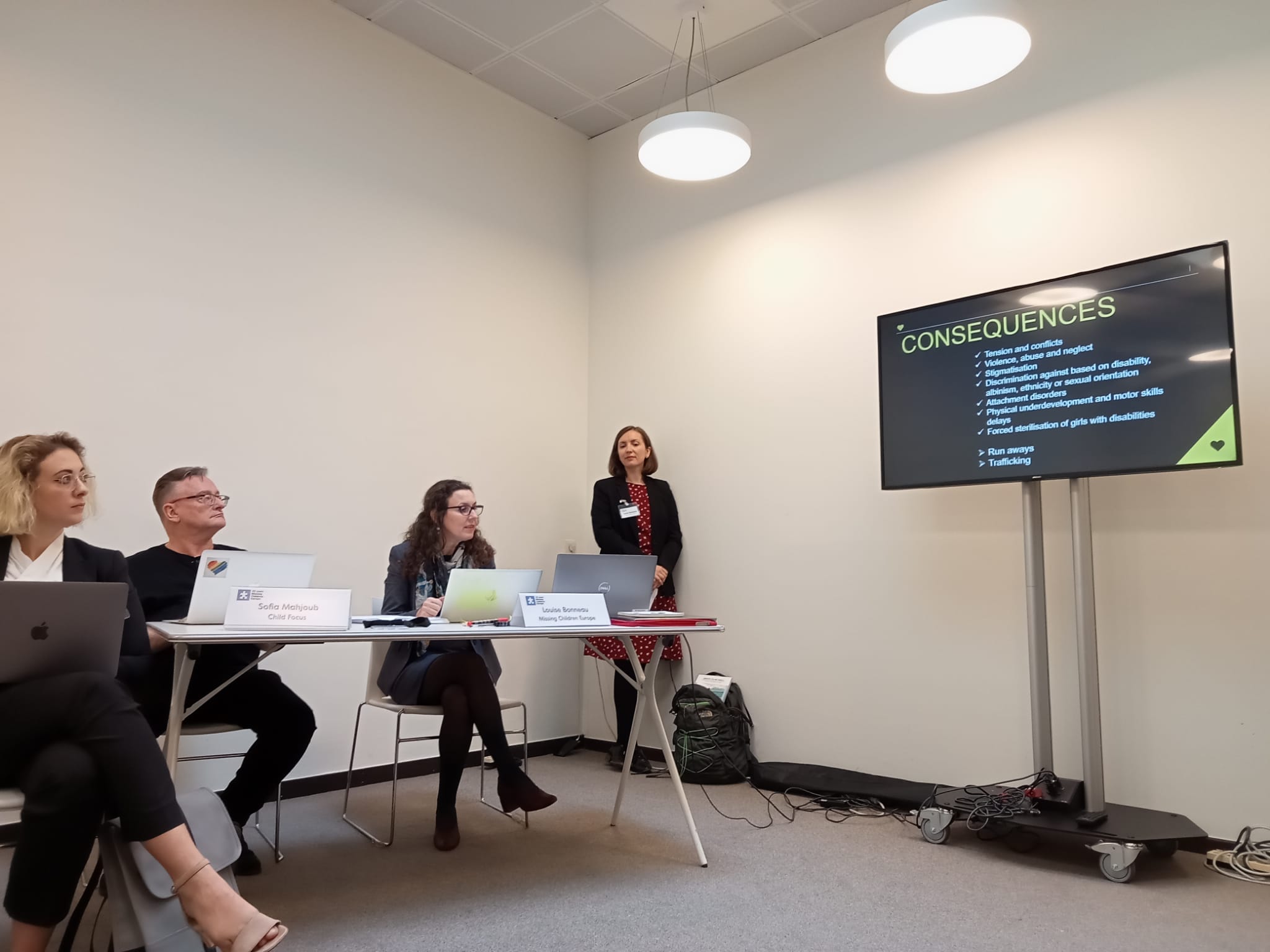 Three professionally dressed men and women sit behind a desk in a plain white office room, while a woman in a suit stands next to a screen on the wall, talking through a presentation.