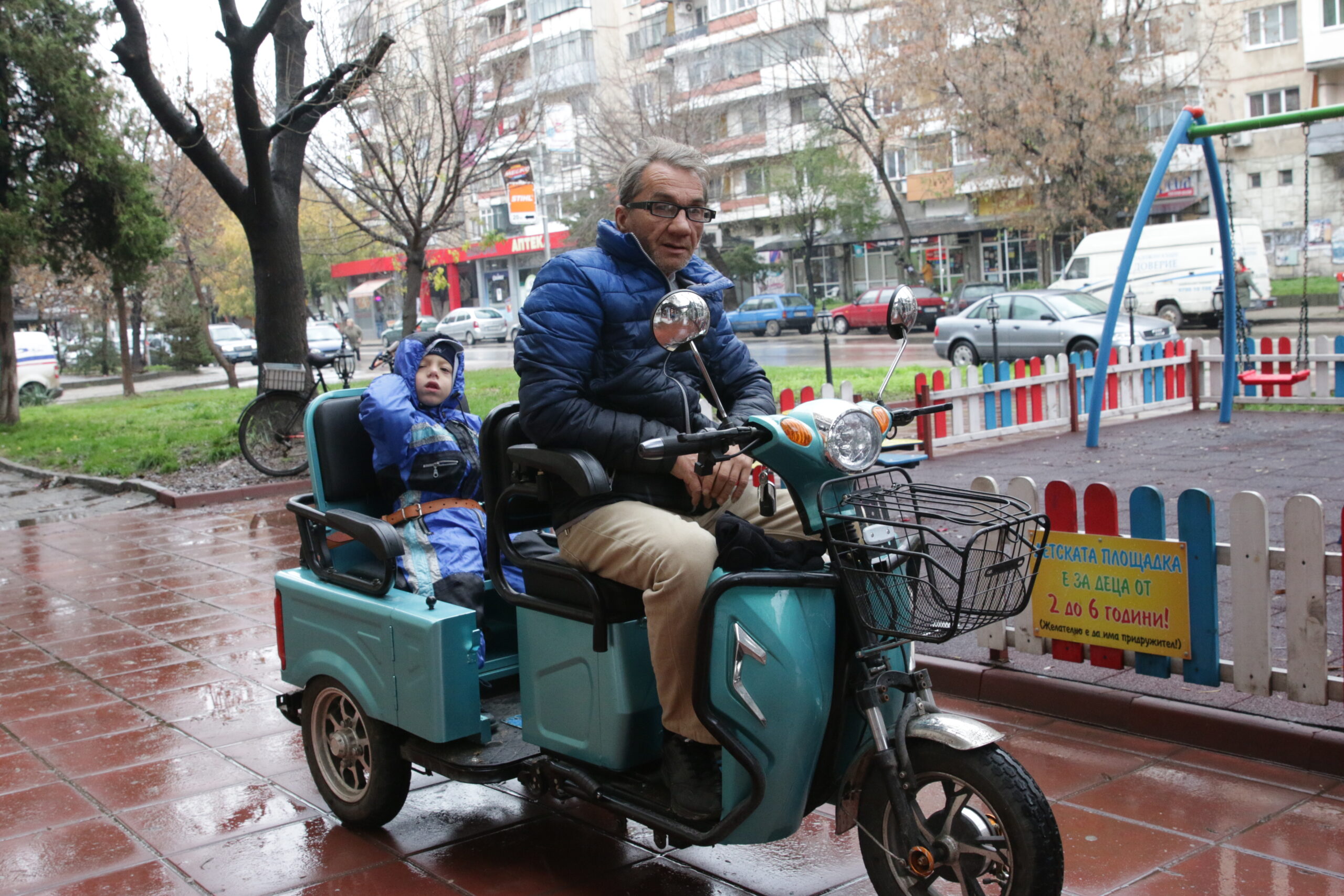 A father and his disabled son ride on a mobility scooter along a tree lined street in the rain