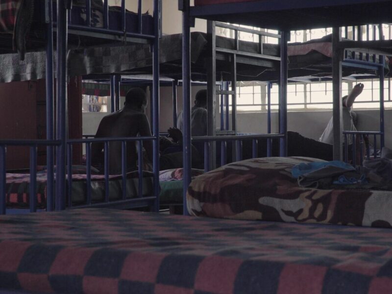 A child sits alone on the bottom bunk of a dim room filled with metal bunk beds, in an orphanage in Kenya