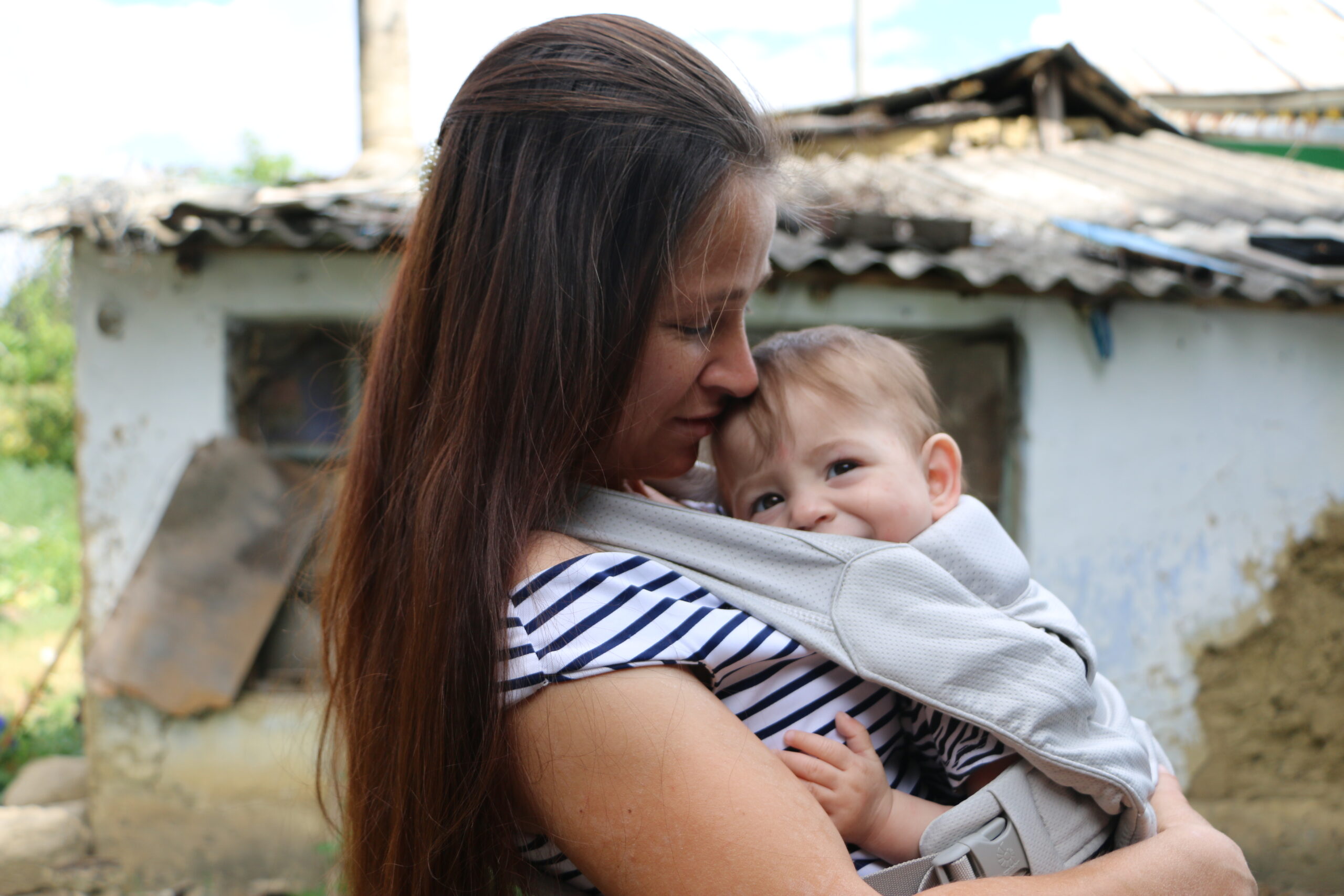 A young Moldovan woman with long dark hair and a stripy top stands with her house in the background, her baby son in a baby carrier on her chest. His head peeps out smiling.