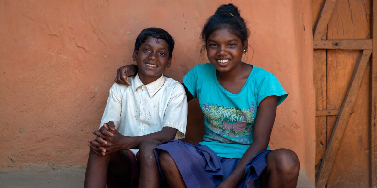 An Indian girl sits with her arm around her younger brother as they both smile at the camera
