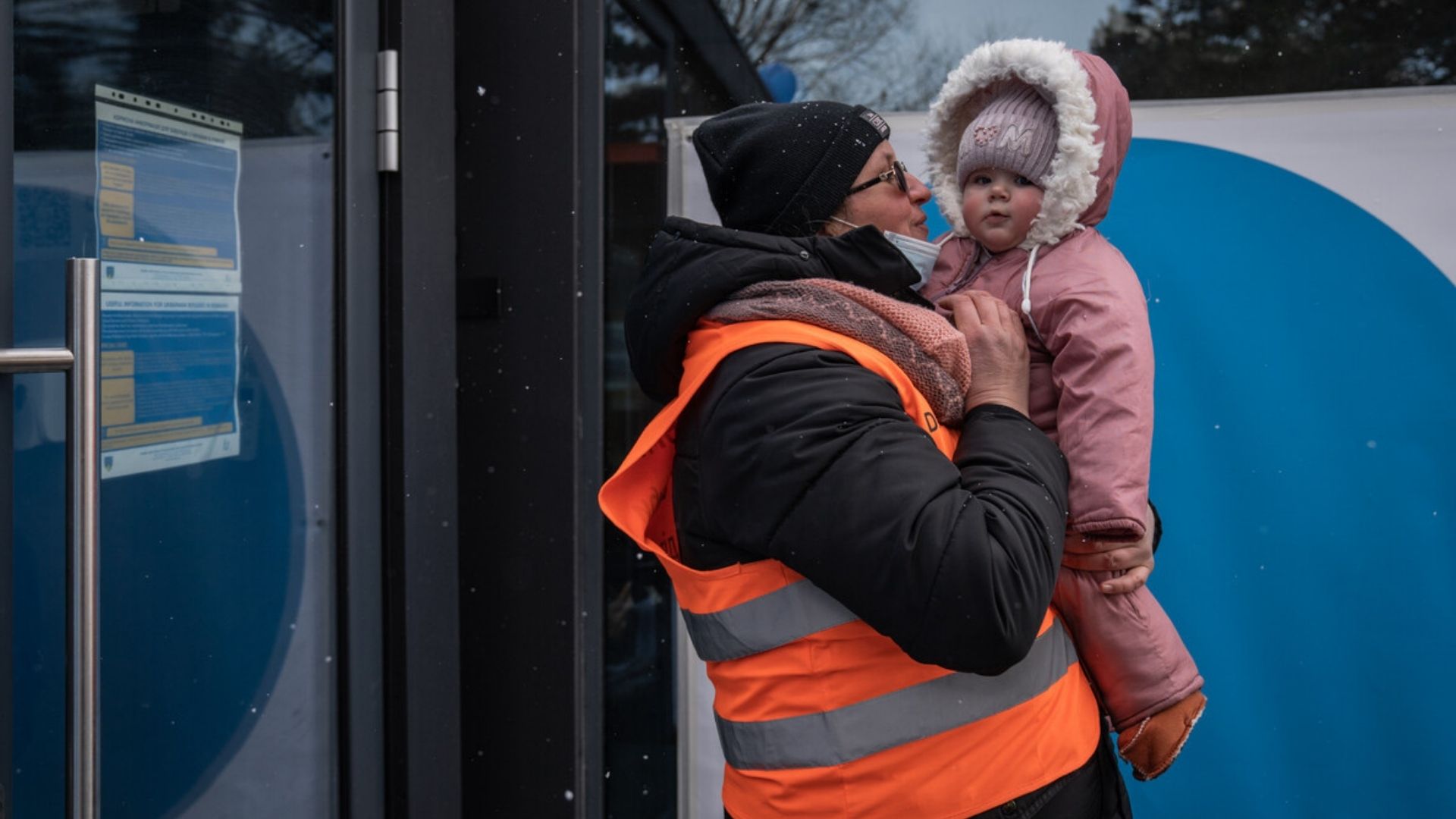 Scenes from the border between Romania and Ukraine during the crisis. A child is being held by a man in a high visibility vest while it snows.