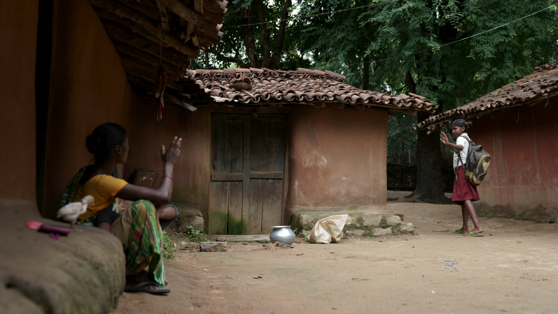 A young indian girl in school uniform waves goodbye to her mother on her way to school outside their rural home