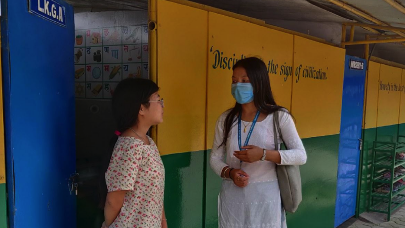 Two Nepalese women chat in front of brightly coloured doors. One wears a mask