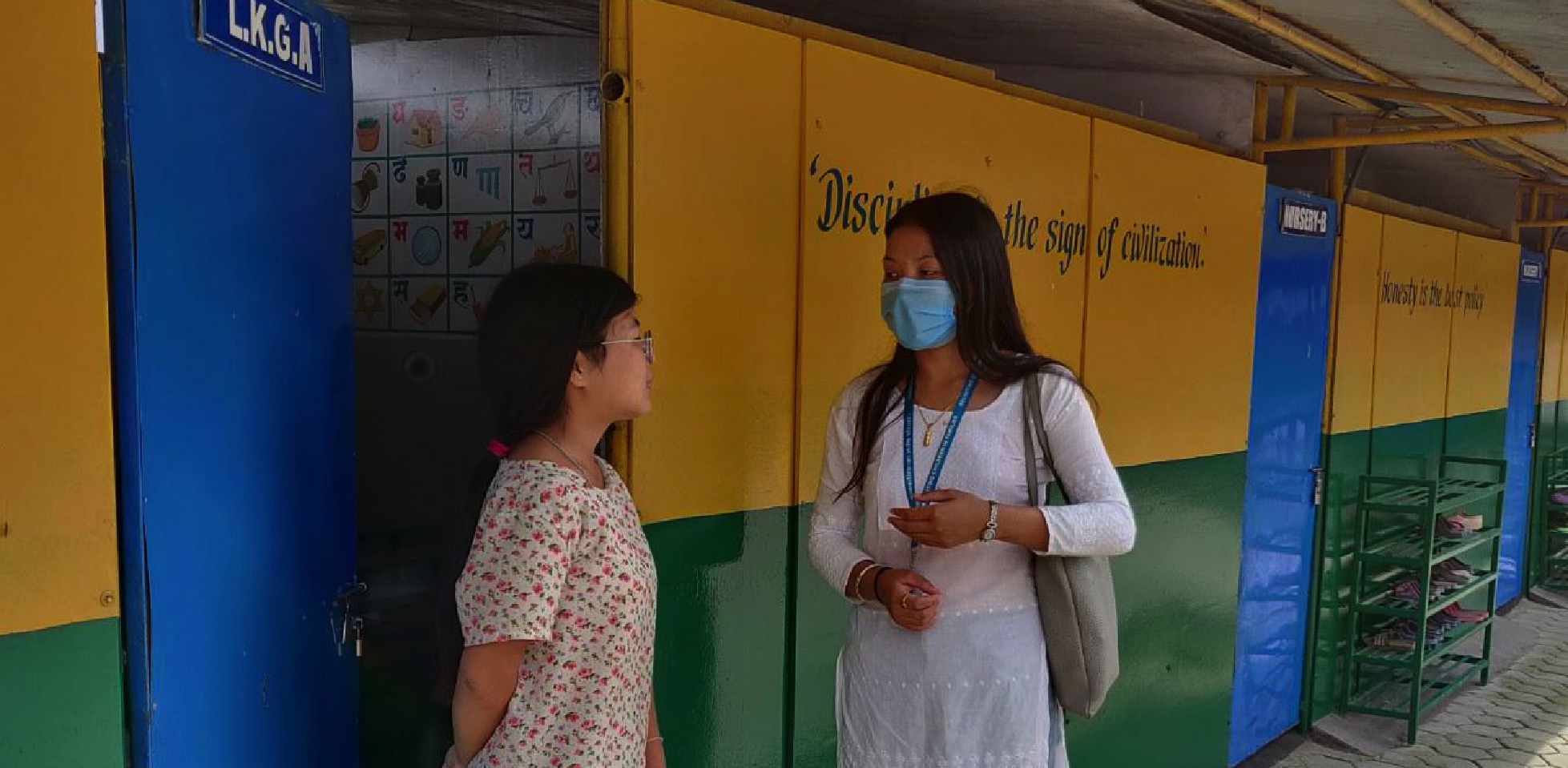 Two Nepalese women chat in front of brightly coloured doors. One wears a mask