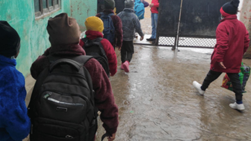 Several warmly dressed Nepali children troop out through an outside door of an orphanage, backs to the camera, wearing rucksacks