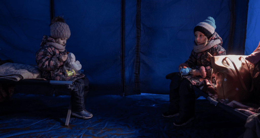 Two children sit in a blue tent on fold out beds. They're wearing beanies, scarves and coats – a symbol of children impacted by displacement on world refugee day.